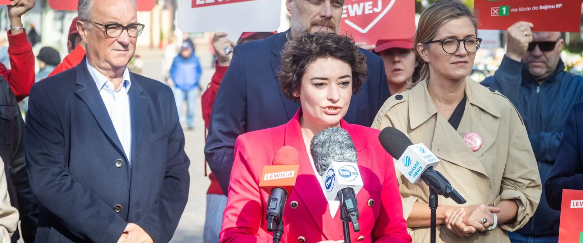 Kobieta w różowym garniturze przemawia do mediów podczas konferencji prasowej Lewicy, otoczona przez zwolenników i plakaty z napisem Lewica.
