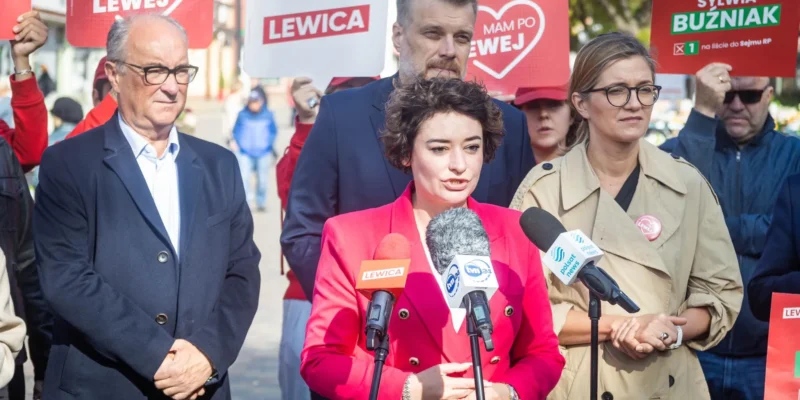 Kobieta w różowym garniturze przemawia do mediów podczas konferencji prasowej Lewicy, otoczona przez zwolenników i plakaty z napisem Lewica.