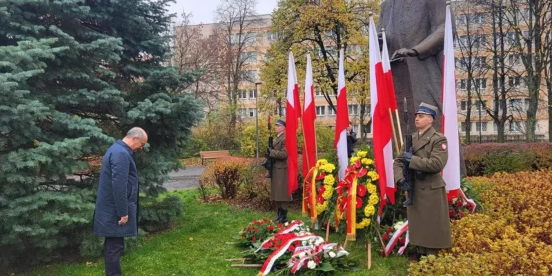 Mężczyzna składa hołd przed pomnikiem podczas obchodów Narodowego Święta Niepodległości, wokół stoją polskie flagi, kwiaty i żołnierze w mundurach.