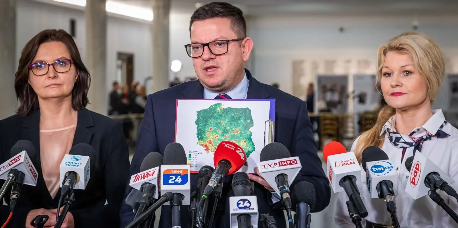 Trzy osoby: dwie kobiety i mężczyzna, stoją na konferencji prasowej; centralnie mężczyzna prezentuje mapę Polski otoczoną mikrofonami.