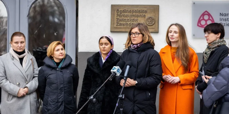Kobiety stoją na zewnątrz budynku podczas konferencji prasowej, z ustawionymi przed nimi mikrofonami, a w tle widoczna tablica z napisem Ogólnopolskie Porozumienie Związków Zawodowych.