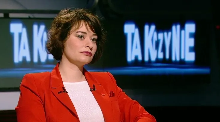 Kobieta o poważnym wyrazie twarzy w czerwonej marynarce siedzi w studiu telewizyjnym podczas programu publicystycznego, z podświetlonym ekranem w tle.