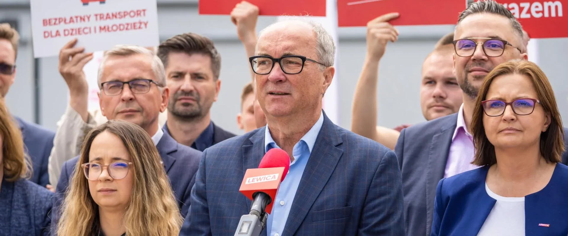 Grupa osób na wiecu Lewicy; mężczyzna w granatowym garniturze przemawia do mikrofonu z czerwonym logiem Lewicy, otoczony przez innych uczestników trzymających plakaty i banery.