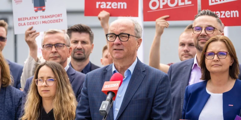 Grupa osób na wiecu Lewicy; mężczyzna w granatowym garniturze przemawia do mikrofonu z czerwonym logiem Lewicy, otoczony przez innych uczestników trzymających plakaty i banery.