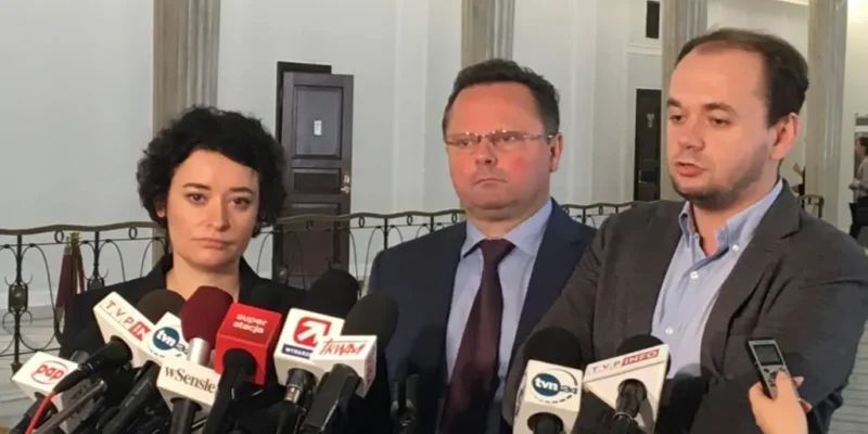 Troje osób podczas konferencji prasowej w korytarzu parlamentu, stojących przed kilkoma mikrofonami i dyktafonami.