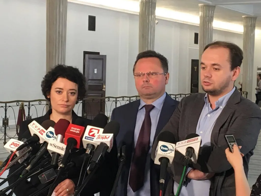 Troje osób podczas konferencji prasowej w korytarzu parlamentu, stojących przed kilkoma mikrofonami i dyktafonami.