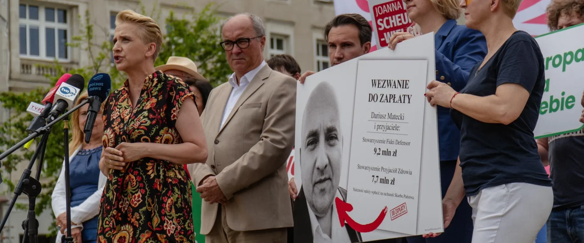 Kobieta przemawia do mikrofonów podczas demonstracji przed budynkiem, a grupa osób trzyma duży plakat z napisem „Wezwanie do zapłaty” i wizerunkiem mężczyzny.