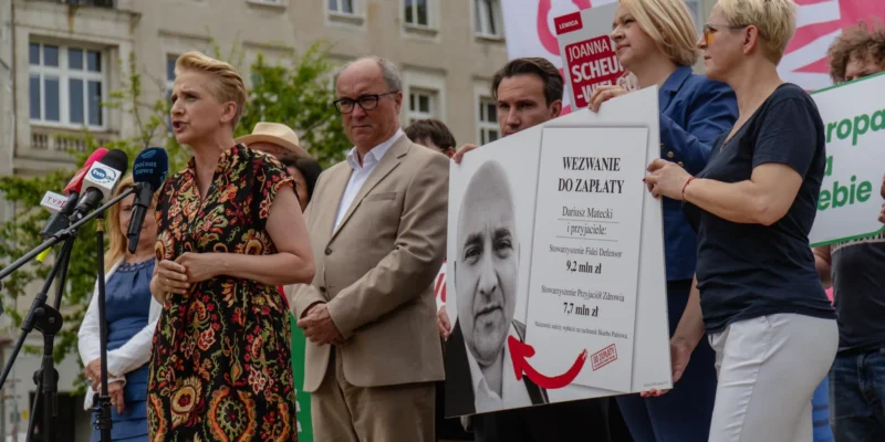Kobieta przemawia do mikrofonów podczas demonstracji przed budynkiem, a grupa osób trzyma duży plakat z napisem „Wezwanie do zapłaty” i wizerunkiem mężczyzny.