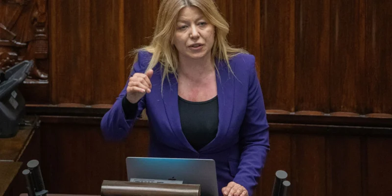 Kobieta o blond włosach w fioletowej marynarce przemawia z mównicy w sali obrad, obok laptopa na biurku.