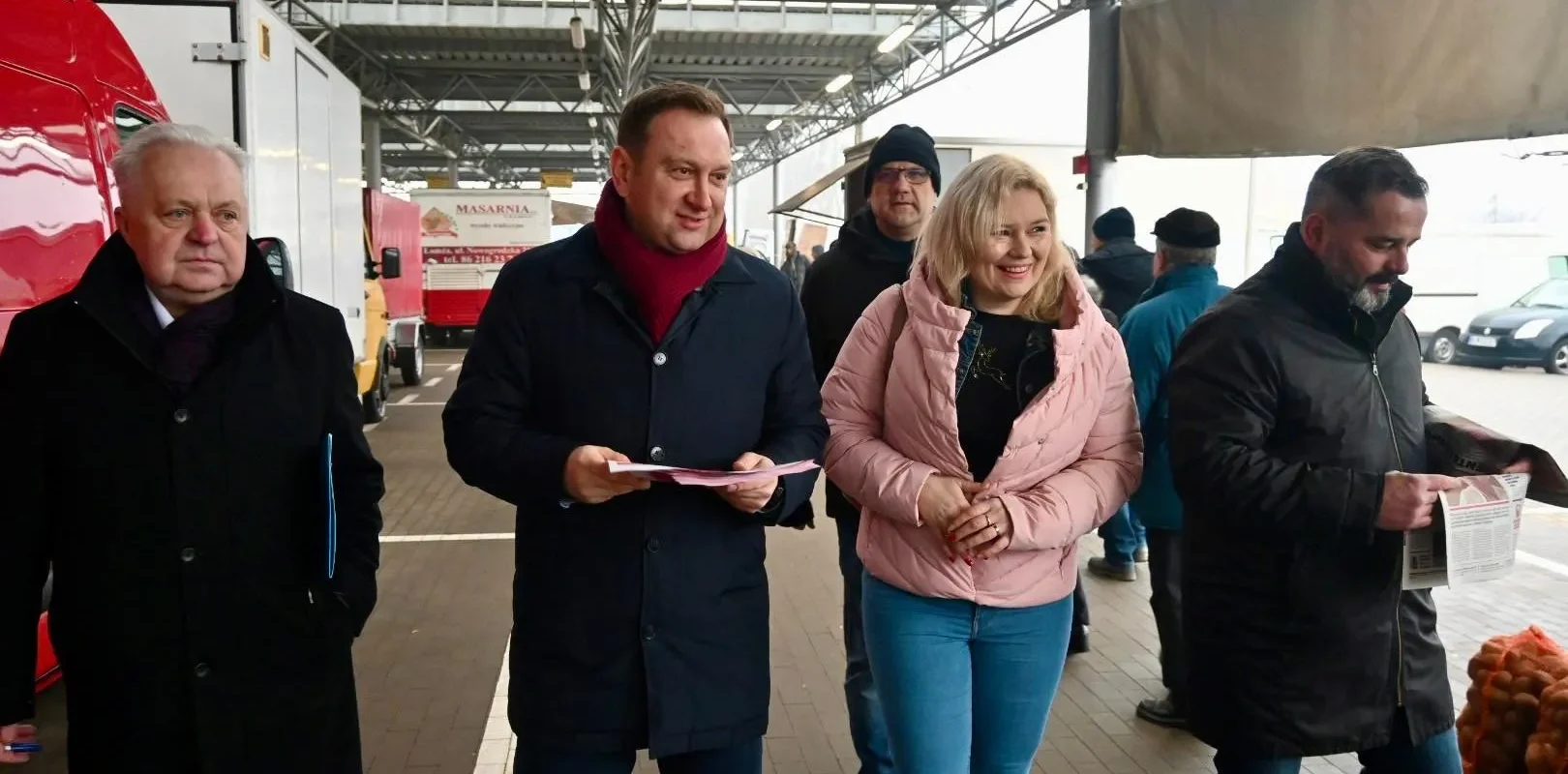 Grupa osób spaceruje po targu, rozmawiając i przeglądając dokumenty, wśród nich dwie uśmiechnięte osoby, mężczyzna w ciemnym płaszczu i kobieta w różowej kurtce.