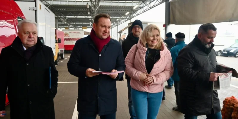 Grupa osób spaceruje po targu, rozmawiając i przeglądając dokumenty, wśród nich dwie uśmiechnięte osoby, mężczyzna w ciemnym płaszczu i kobieta w różowej kurtce.