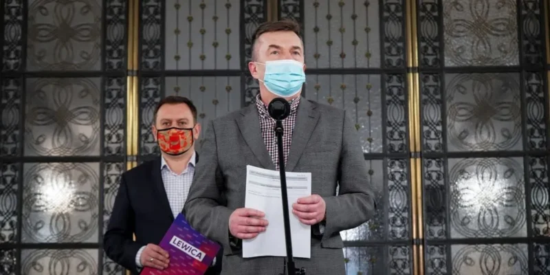 Dwóch mężczyzn w garniturach i maseczkach stoi przy mikrofonie podczas konferencji prasowej; pierwszy trzyma kartki, a za nim drugi trzyma plik z napisem Lewica na tle ozdobnych przeszklonych drzwi.