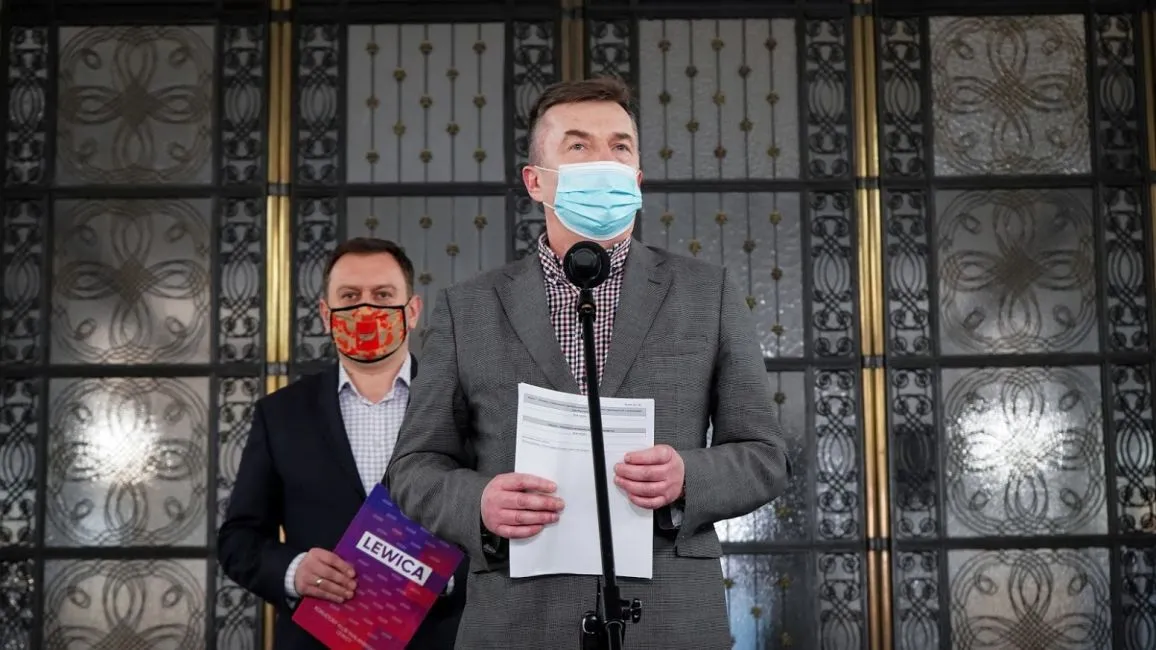 Dwóch mężczyzn w garniturach i maseczkach stoi przy mikrofonie podczas konferencji prasowej; pierwszy trzyma kartki, a za nim drugi trzyma plik z napisem Lewica na tle ozdobnych przeszklonych drzwi.