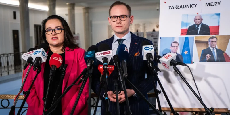 Dwóch polityków stoi przy zestawie mikrofonów podczas konferencji prasowej, obok planszy z fotografiami i napisem „ZAKŁADNICY MEJZY”.