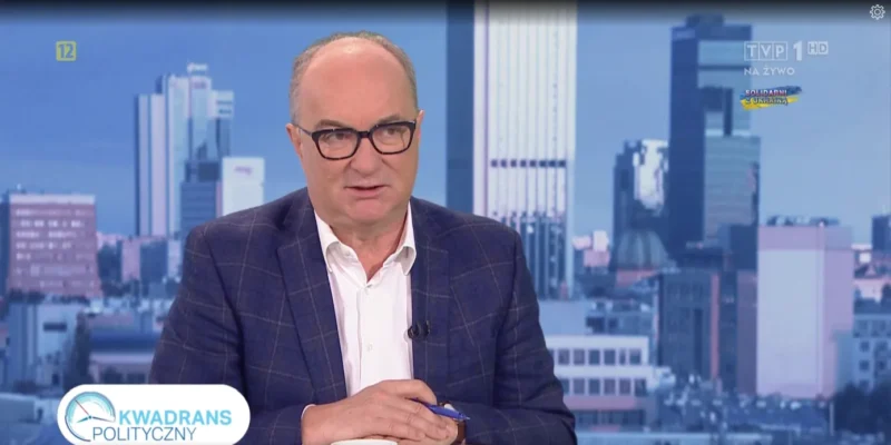 Na zdjęciu widoczny jest mężczyzna w okularach, ubrany w ciemną marynarkę i białą koszulę, który siedzi przy biurku w studiu telewizyjnym z tłem miejskim.