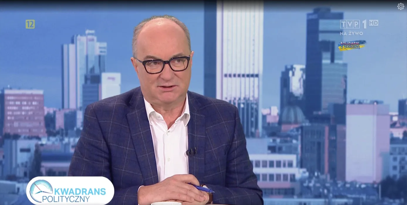 Na zdjęciu widoczny jest mężczyzna w okularach, ubrany w ciemną marynarkę i białą koszulę, który siedzi przy biurku w studiu telewizyjnym z tłem miejskim.