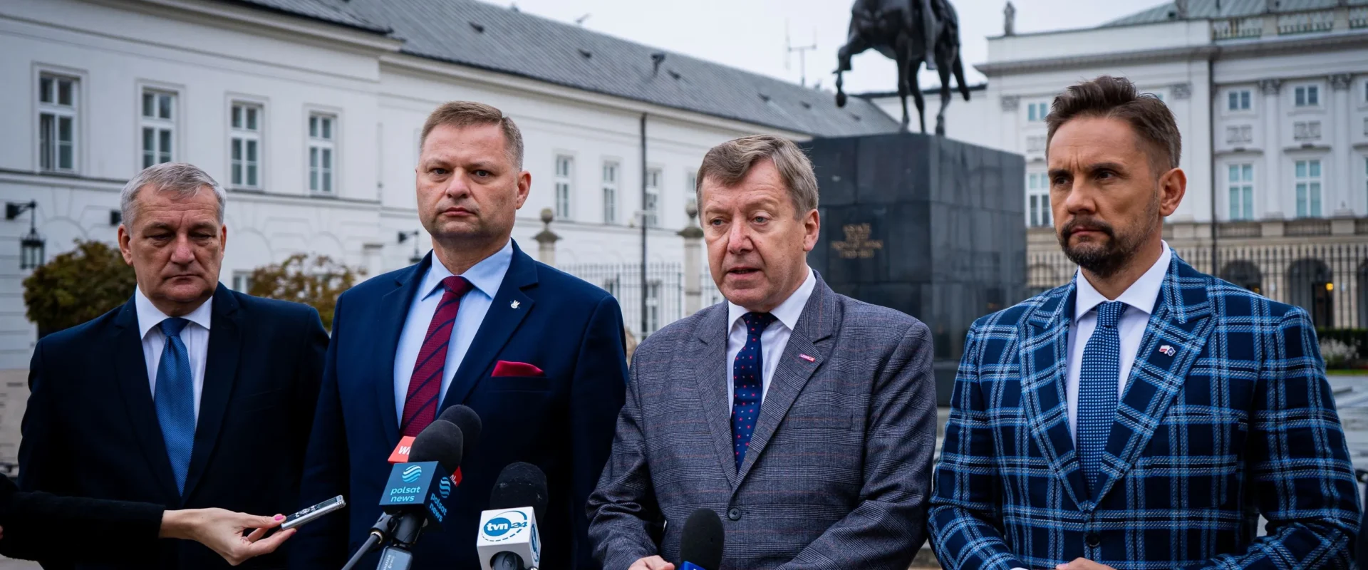 Czterech mężczyzn w garniturach podczas konferencji prasowej stojących przed pomnikiem jeźdźca i budynkami urzędowymi; w dole widać mikrofony dziennikarskie.