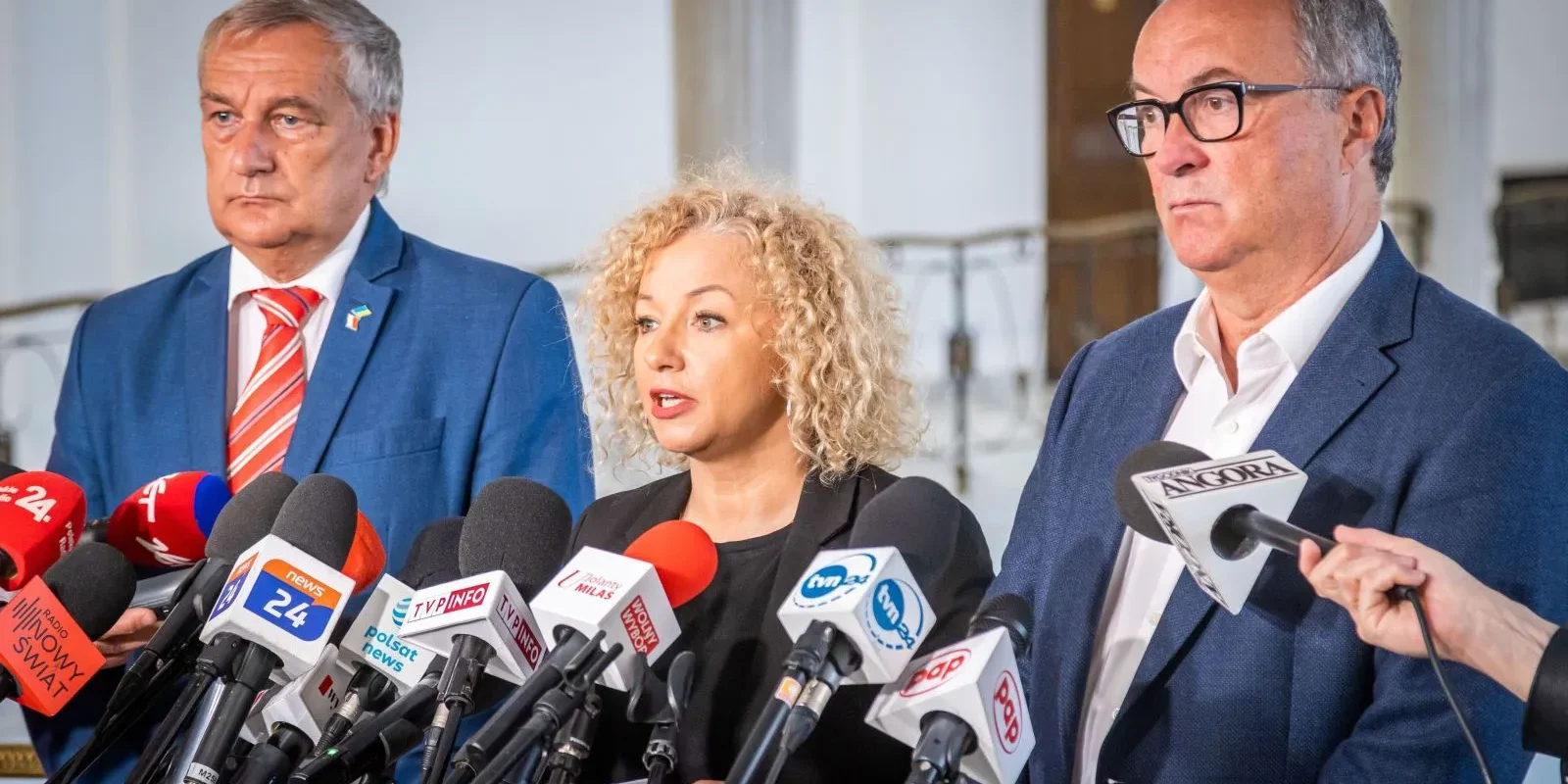 Na zdjęciu trzej przedstawiciele mówią do zgromadzonych dziennikarzy przed mikrofonami w oficjalnym otoczeniu.