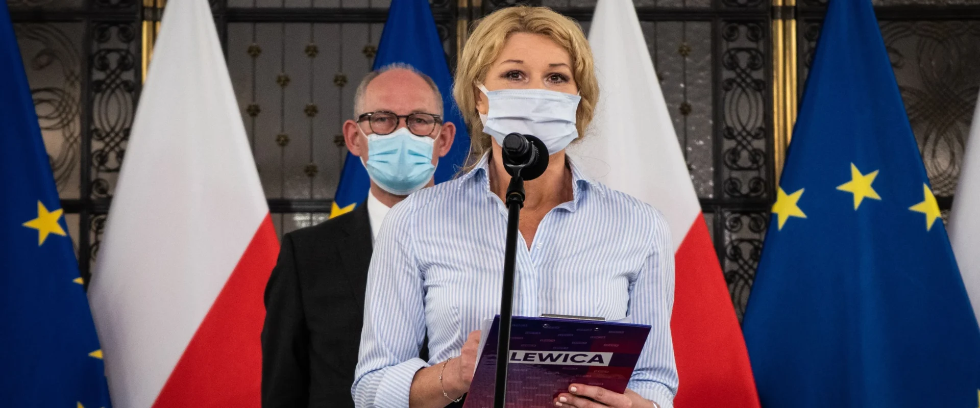 Kobieta w maseczce stoi przy mikrofonie podczas konferencji prasowej, trzyma notes z napisem LEWICA, a w tle widać flagi Polski i Unii Europejskiej.