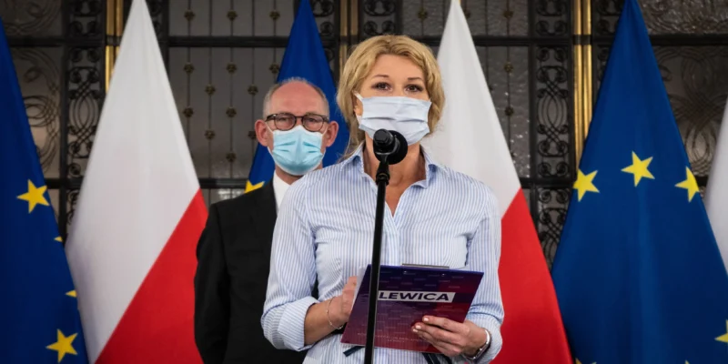 Kobieta w maseczce stoi przy mikrofonie podczas konferencji prasowej, trzyma notes z napisem LEWICA, a w tle widać flagi Polski i Unii Europejskiej.