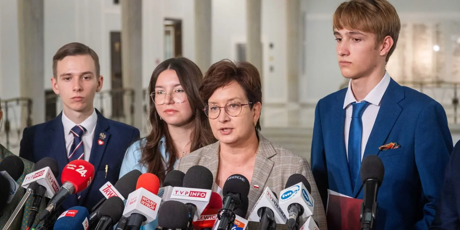 Kobieta w okularach przemawia do licznych mikrofonów podczas konferencji prasowej, otoczona przez młodych ludzi w garniturach.