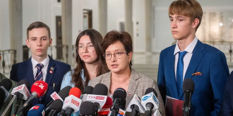 Kobieta w okularach przemawia do licznych mikrofonów podczas konferencji prasowej, otoczona przez młodych ludzi w garniturach.