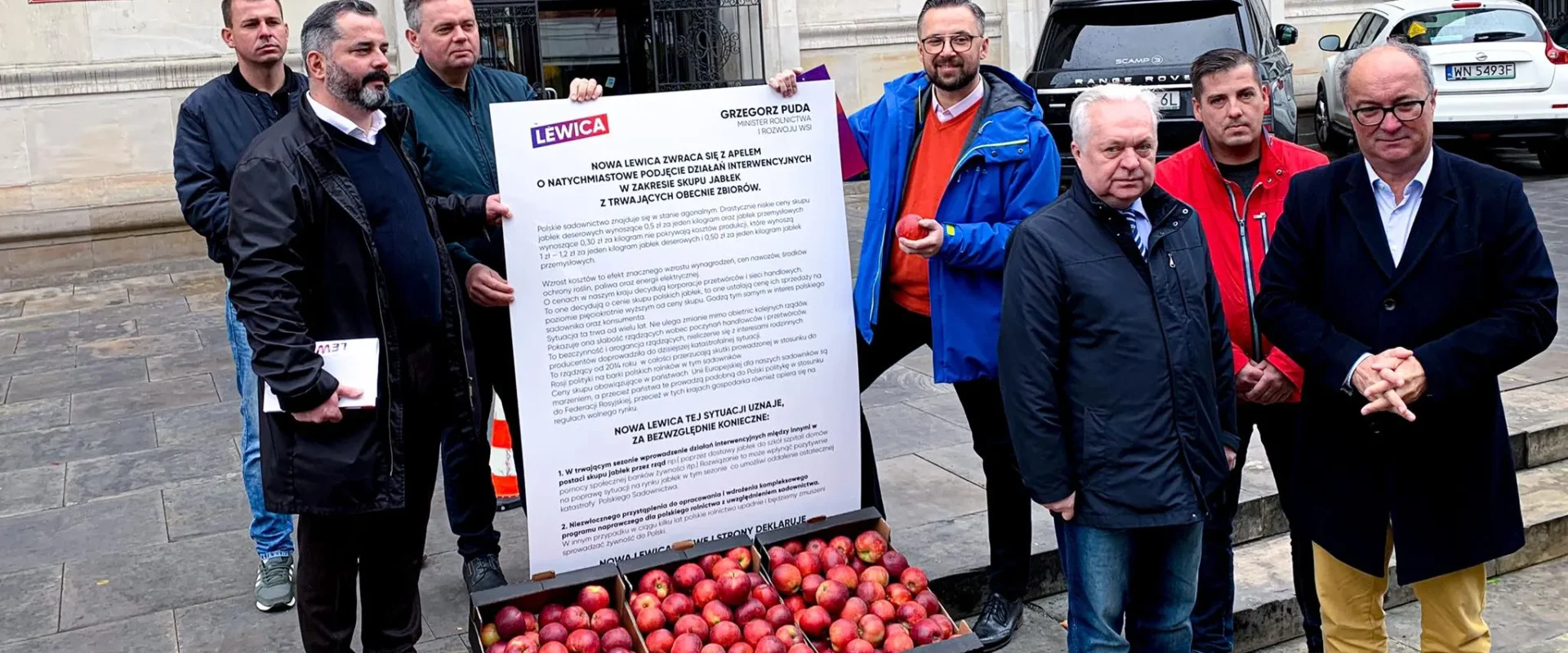 Grupa mężczyzn stojących przed wejściem do budynku Ministerstwa Rolnictwa i Rozwoju Wsi, trzymających duży plakat Lewicy i skrzynie z jabłkami ustawione na schodach.