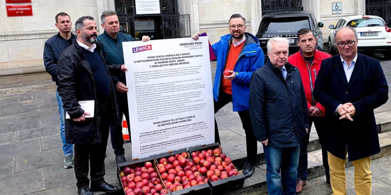 Grupa mężczyzn stojących przed wejściem do budynku Ministerstwa Rolnictwa i Rozwoju Wsi, trzymających duży plakat Lewicy i skrzynie z jabłkami ustawione na schodach.