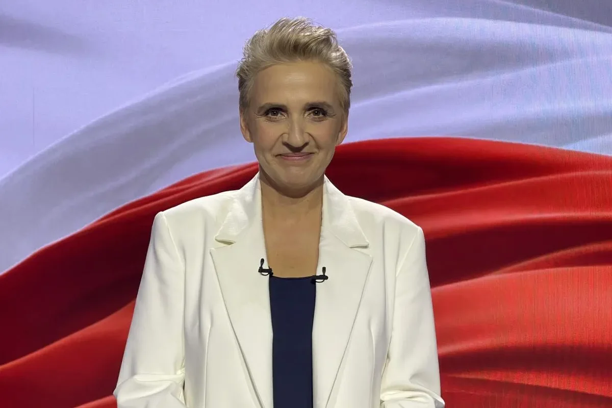 Prowadząca program w białym żakiecie na tle czerwono-białego studia TVP, z mikrofonami na klipsach.