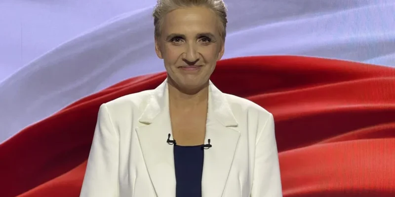 Prowadząca program w białym żakiecie na tle czerwono-białego studia TVP, z mikrofonami na klipsach.