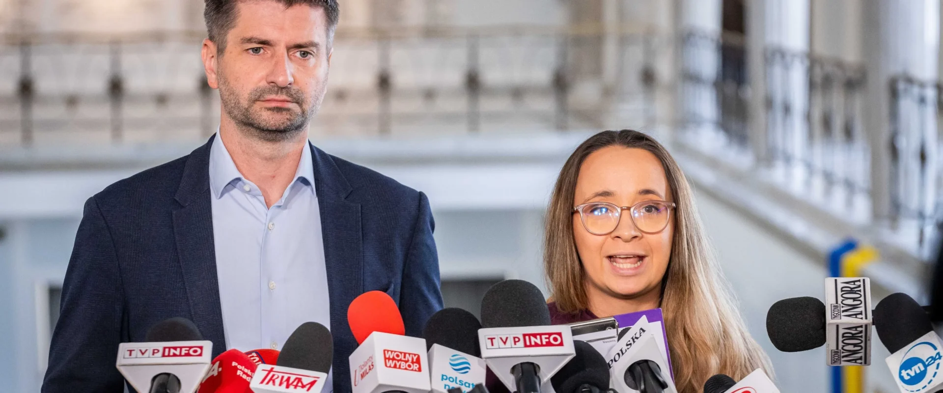 Dwóch mówców stoi przy licznym zestawie mikrofonów podczas konferencji prasowej w eleganckim wnętrzu budynku.