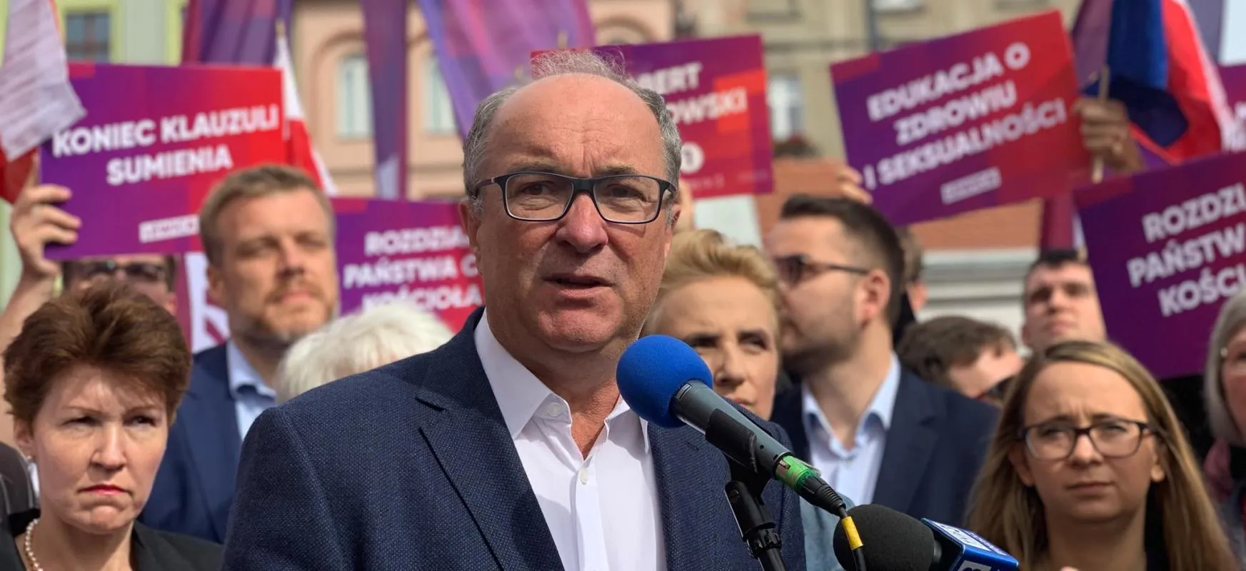 Mężczyzna w okularach w garniturze przemawia do mikrofonu podczas wiecu politycznego opozycji, z plakatami w tle.