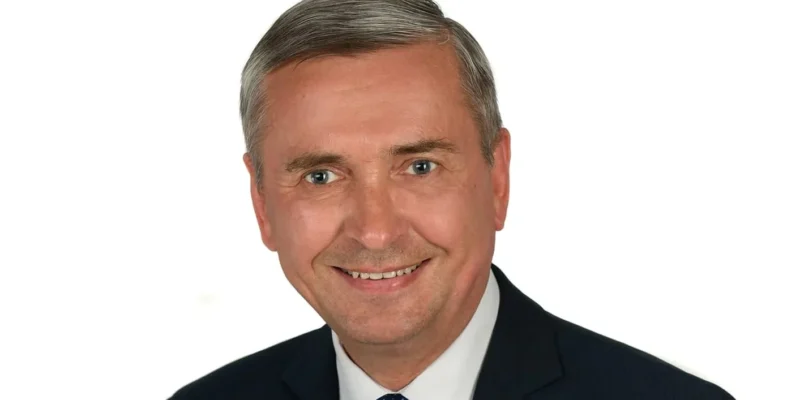 Portret mężczyzny w garniturze i niebieskim krawacie w białe kropki na białym tle, uśmiechnięty.