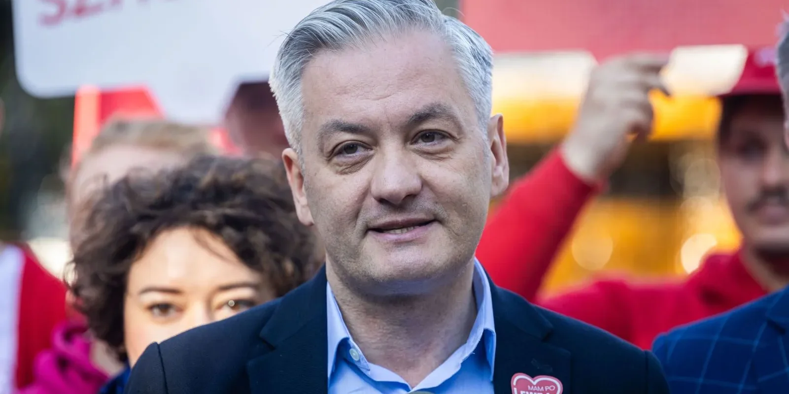 Mężczyzna w granatowej marynarce i jasnoniebieskiej koszuli stoi na tle tłumu podczas wydarzenia, z sercową przypinką na klapie i widocznym mikrofonem w dolnej części kadru.