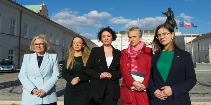 Pięć posłanek Lewicy stoi na placu przed budynkiem z posągiem i flagami, ubranych w eleganckie marynarki, podczas wydarzenia w sprawie ustawy o tabletce „dzień po”.