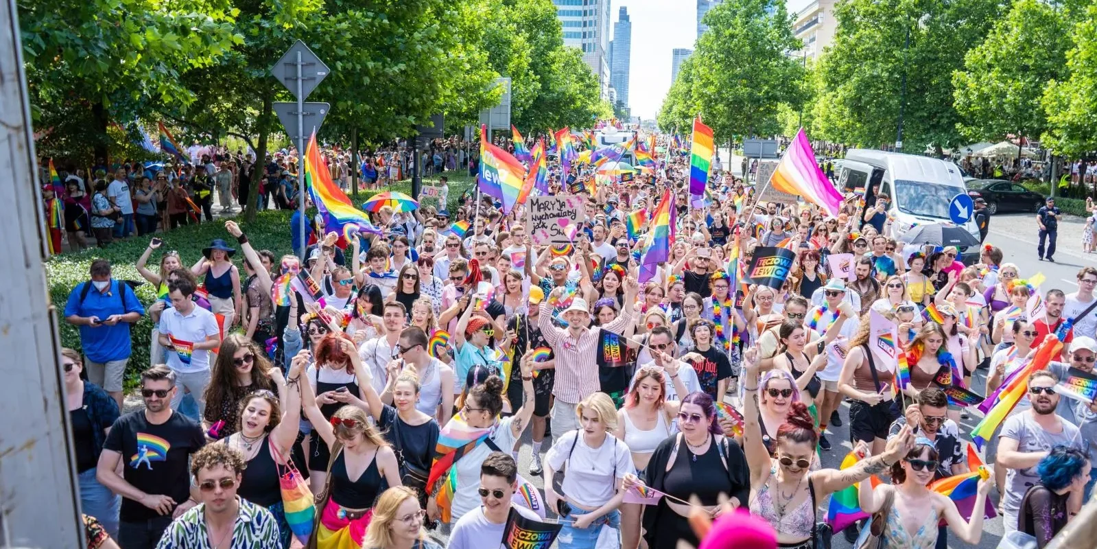 Uliczna scena Parady Równości 2022: tłum ludzi maszeruje, trzymając tęczowe flagi i transparenty w słoneczny dzień pośród zieleni i miejskich budynków.