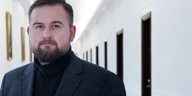 Mężczyzna z krótkimi włosami i brodą, ubrany w ciemny garnitur i czarny golf, stoi w jasnym korytarzu z obrazami na ścianach.