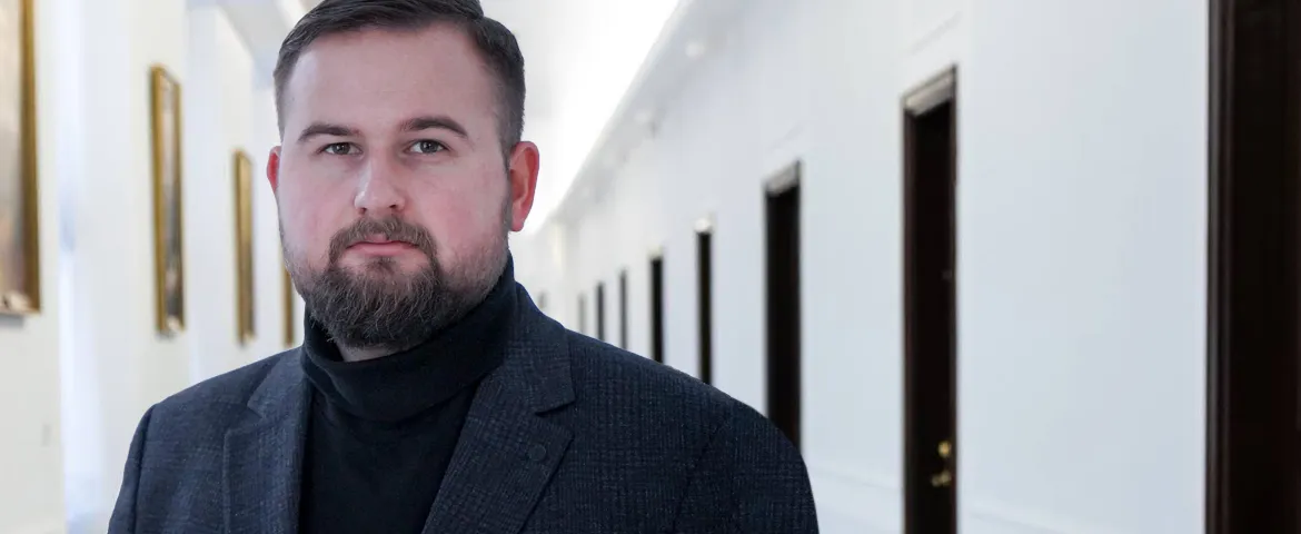Mężczyzna z krótkimi włosami i brodą, ubrany w ciemny garnitur i czarny golf, stoi w jasnym korytarzu z obrazami na ścianach.