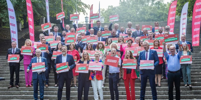 Grupa kandydatów Lewicy na jedynki do Sejmu i Senatu stoi na schodach, trzymając kolorowe plakaty z nazwiskami i logiem Lewicy.