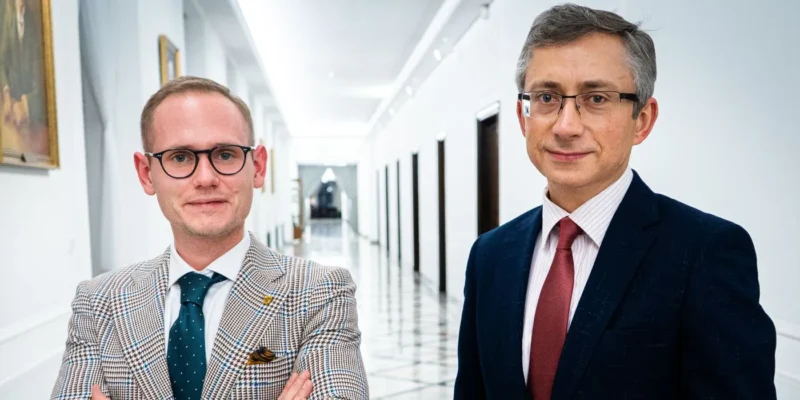 Dwóch mężczyzn w garniturach stoi w jasnym, długim korytarzu z obrazami na ścianach.