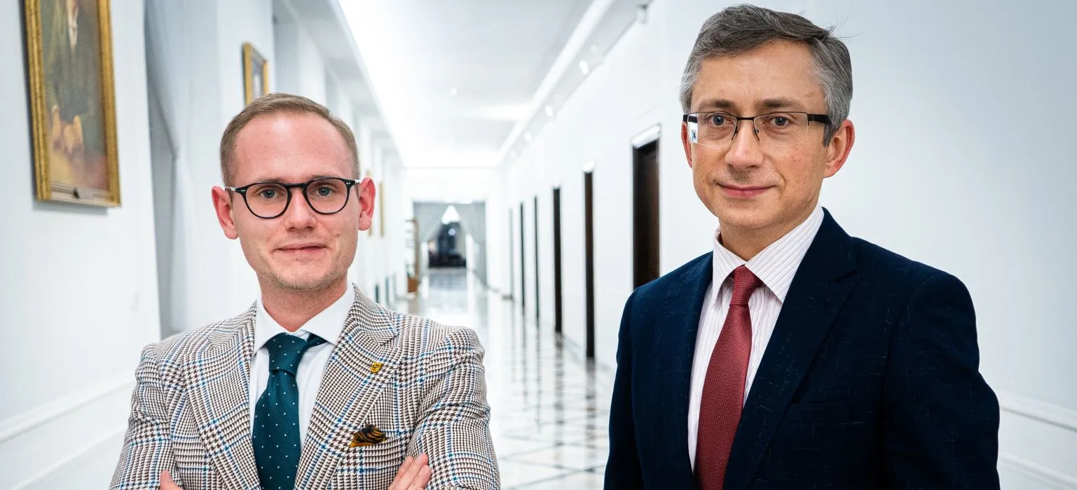 Dwóch mężczyzn w garniturach stoi w jasnym, długim korytarzu z obrazami na ścianach.