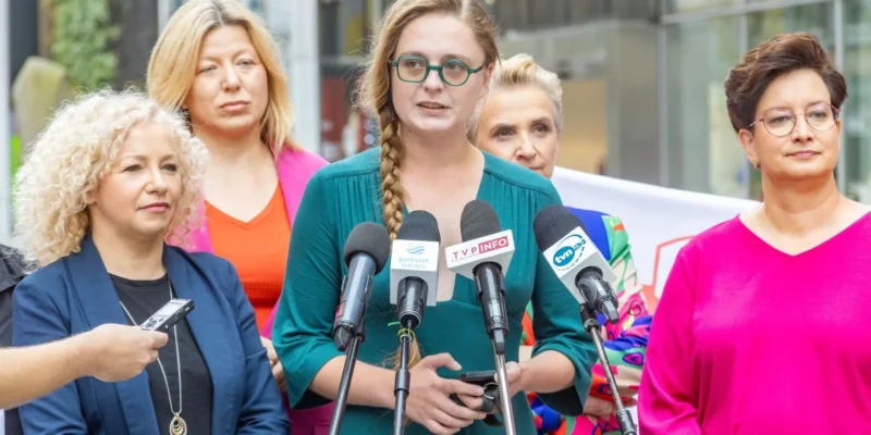 Kobieta w zielonej bluzce przemawia do mediów podczas konferencji prasowej Lewicy, otoczona innymi kobietami.
