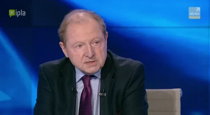 Mężczyzna w garniturze i krawacie siedzi w studiu telewizyjnym na niebieskim tle podczas programu o polityce wschodniej.