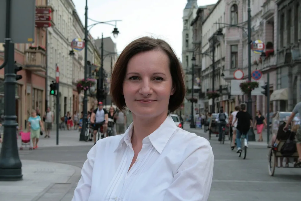 Kobieta w białej koszuli stoi na ruchliwym deptaku miejskim, a w tle widać przechodniów, rowerzystów i kamienice.