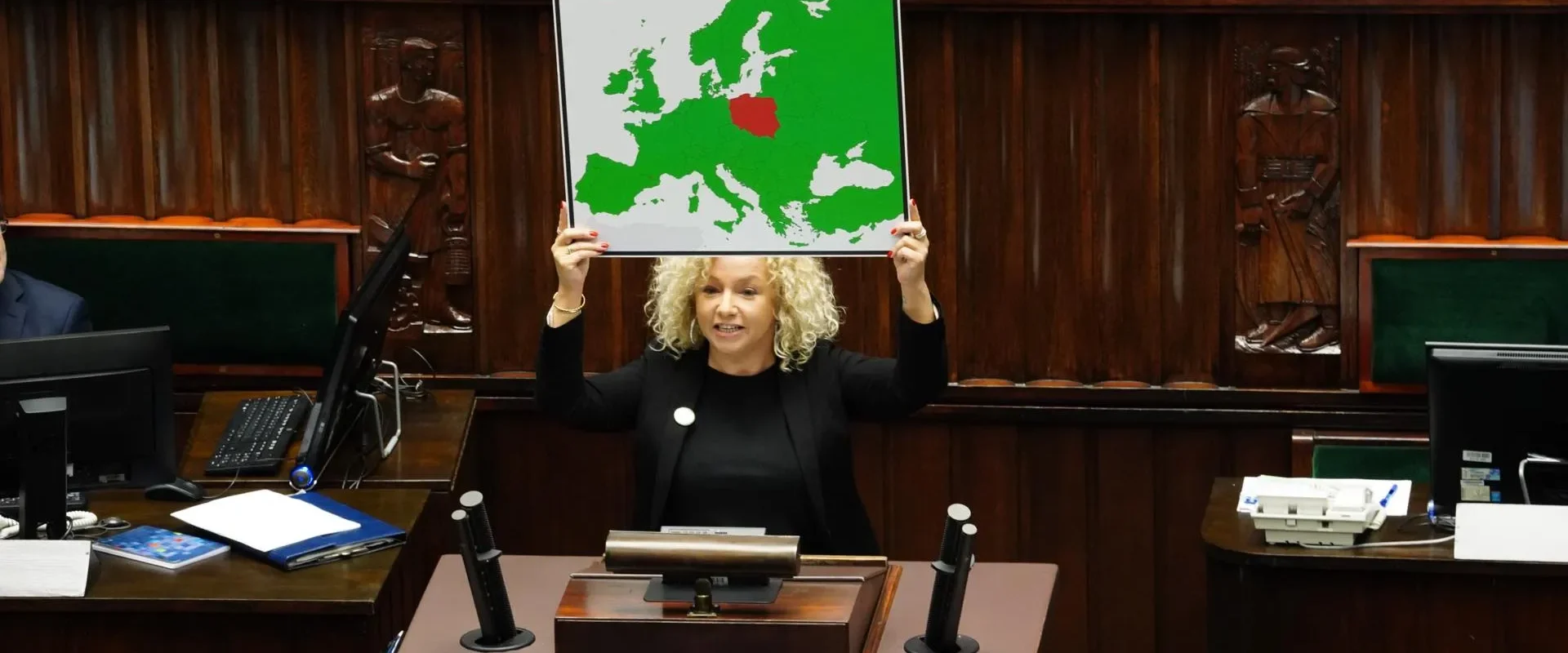 Na zdjęciu widać kobietę z kręconymi włosami, trzymającą mapę Europy, na której czerwono zaznaczone jest jedno z państw, w tle widać salę obrad.