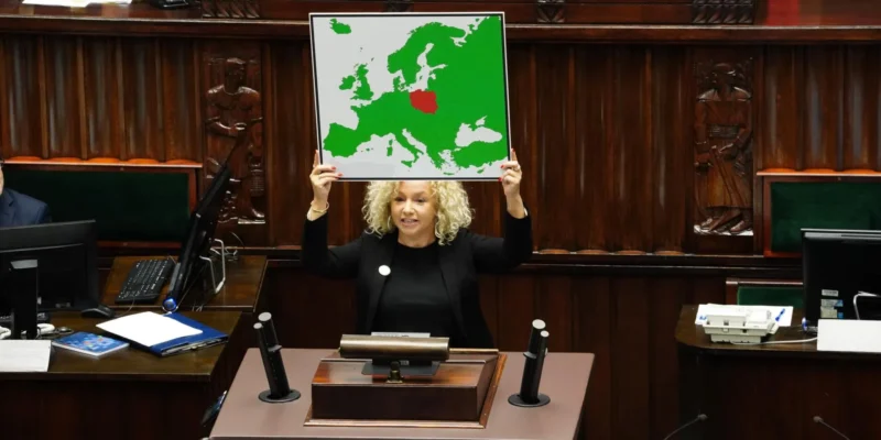 Na zdjęciu widać kobietę z kręconymi włosami, trzymającą mapę Europy, na której czerwono zaznaczone jest jedno z państw, w tle widać salę obrad.