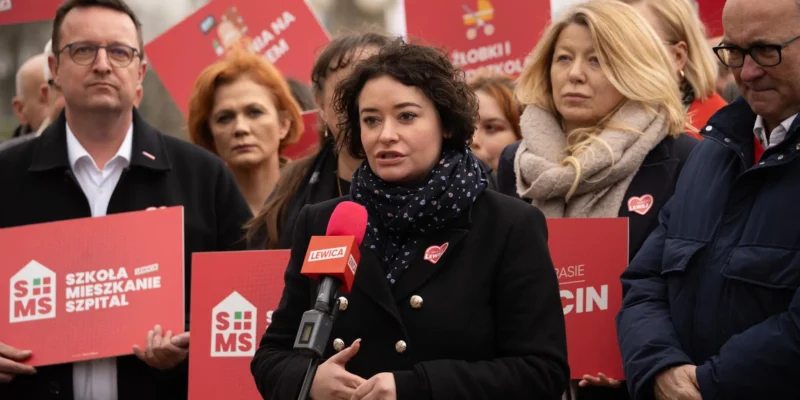 Kobieta przemawia na wiecu Lewicy, otoczona innymi uczestnikami trzymającymi czerwone plakaty z hasłami i logo partii.