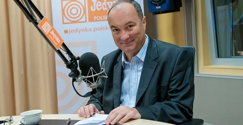 Mężczyzna w marynarce uśmiecha się, siedząc w studio radiowym przy mikrofonie, z notatkami i filiżanką na stole oraz widocznym logo stacji w tle.