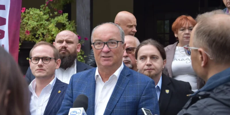 Na zdjęciu grupa mężczyzn i kobiet stoi na zewnątrz, a mężczyzna w niebieskiej marynarce mówi do mikrofonu w otoczeniu innych, z tłem roślinności i baneru.