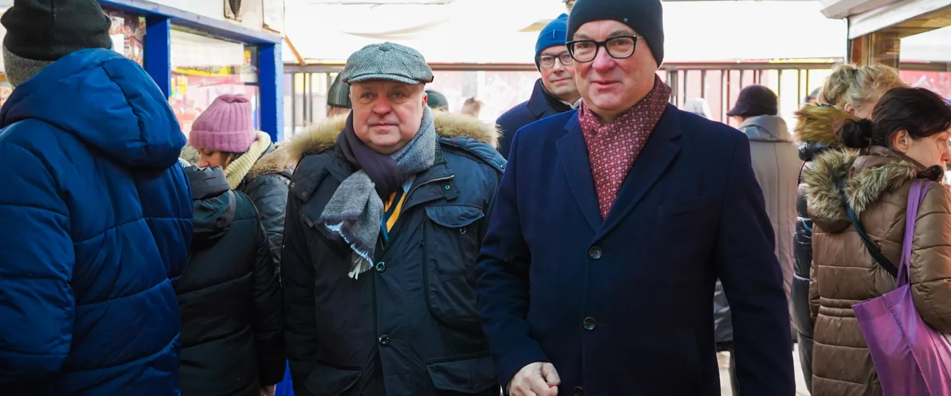 Dwoje mężczyzn w zimowych płaszczach i czapkach spaceruje po targu, otoczeni innymi kupującymi.